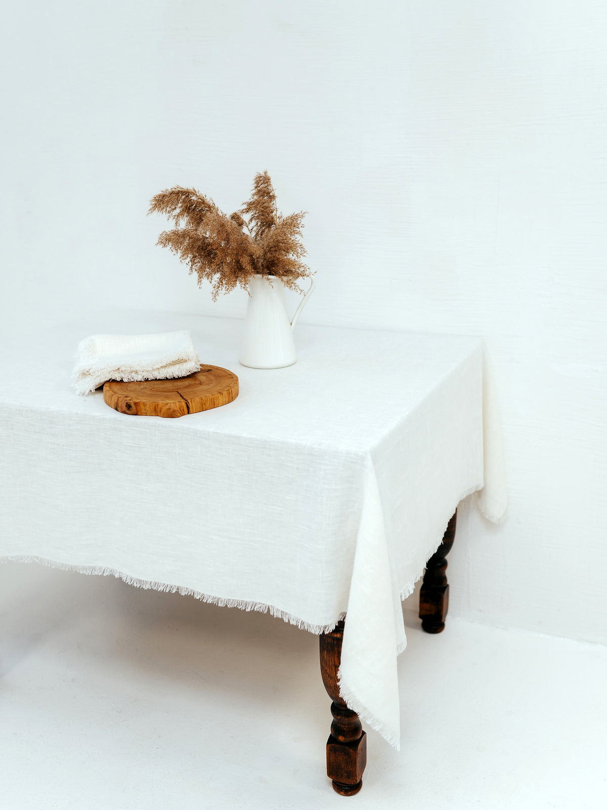 White linen tablecloth covering an antique wooden table. Frayed edge napkins, also in white, are on the table along with a wooden dish and a vase holding dry grasses. 