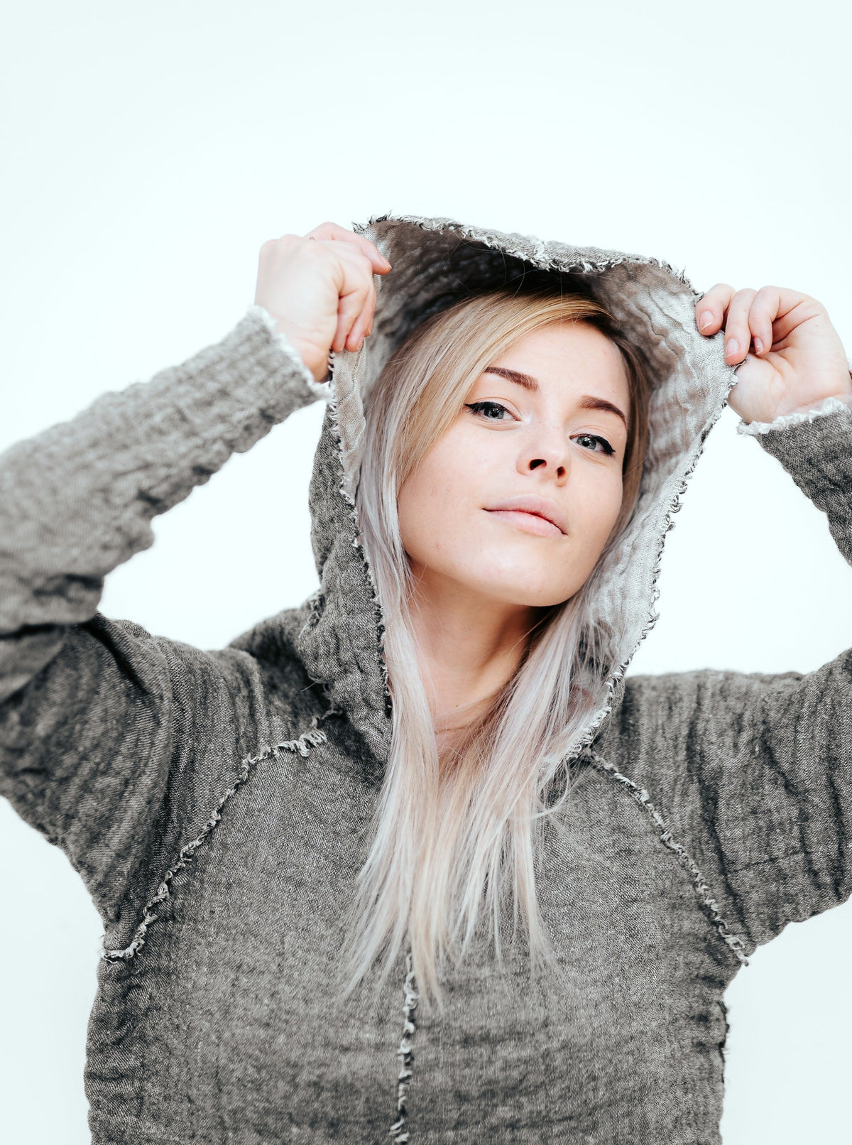 Woman wearing a grey linen and wool pullover with a hood. Her arms are raised and she is lifting the hood while looking into the camera